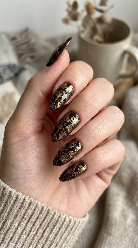 Long almond nails with liquid gold ginkgo leaf illustration on dark chocolate brown gel