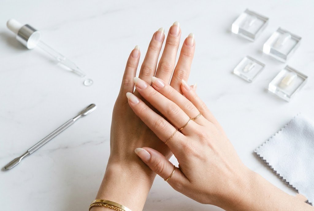 Woman's hands with bare natural nails on marble surface surrounded by nail serum dropper and tools showing nail strengthening routine